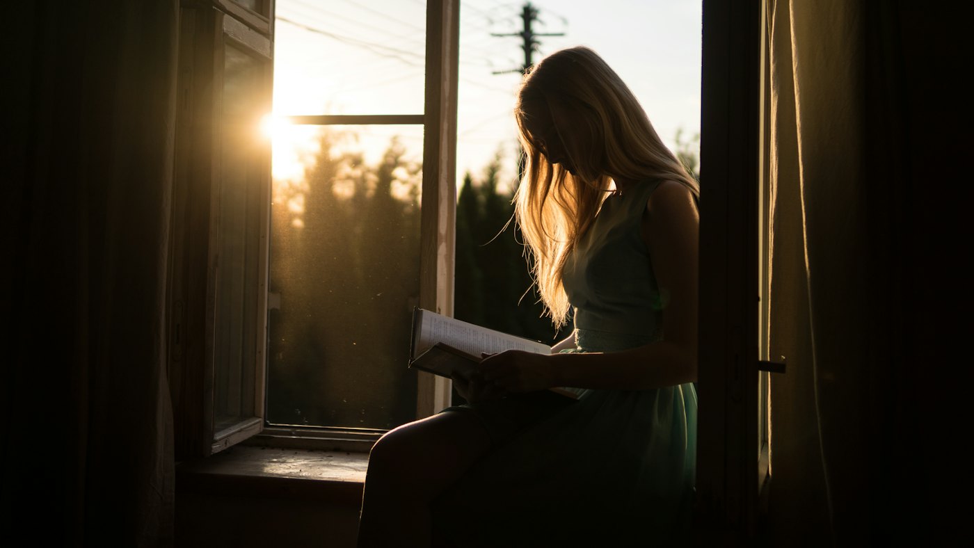 A woman reading by an open window in evening light